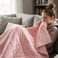 A woman sits on a gray couch in a living room, wrapped in a soft pink weightedblanket with a small embroidered pumpkin design visible on the corner.