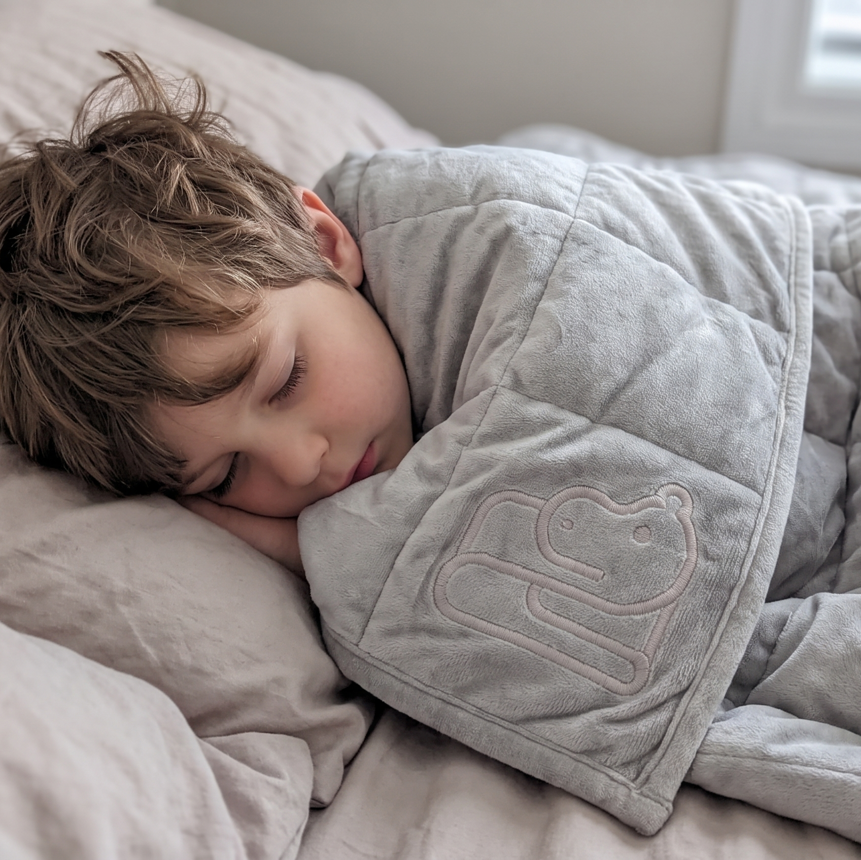 A young child sleeping soundly on a bed, wrapped in a soft, light gray weighted blanket with a stitched hippo design on the front.