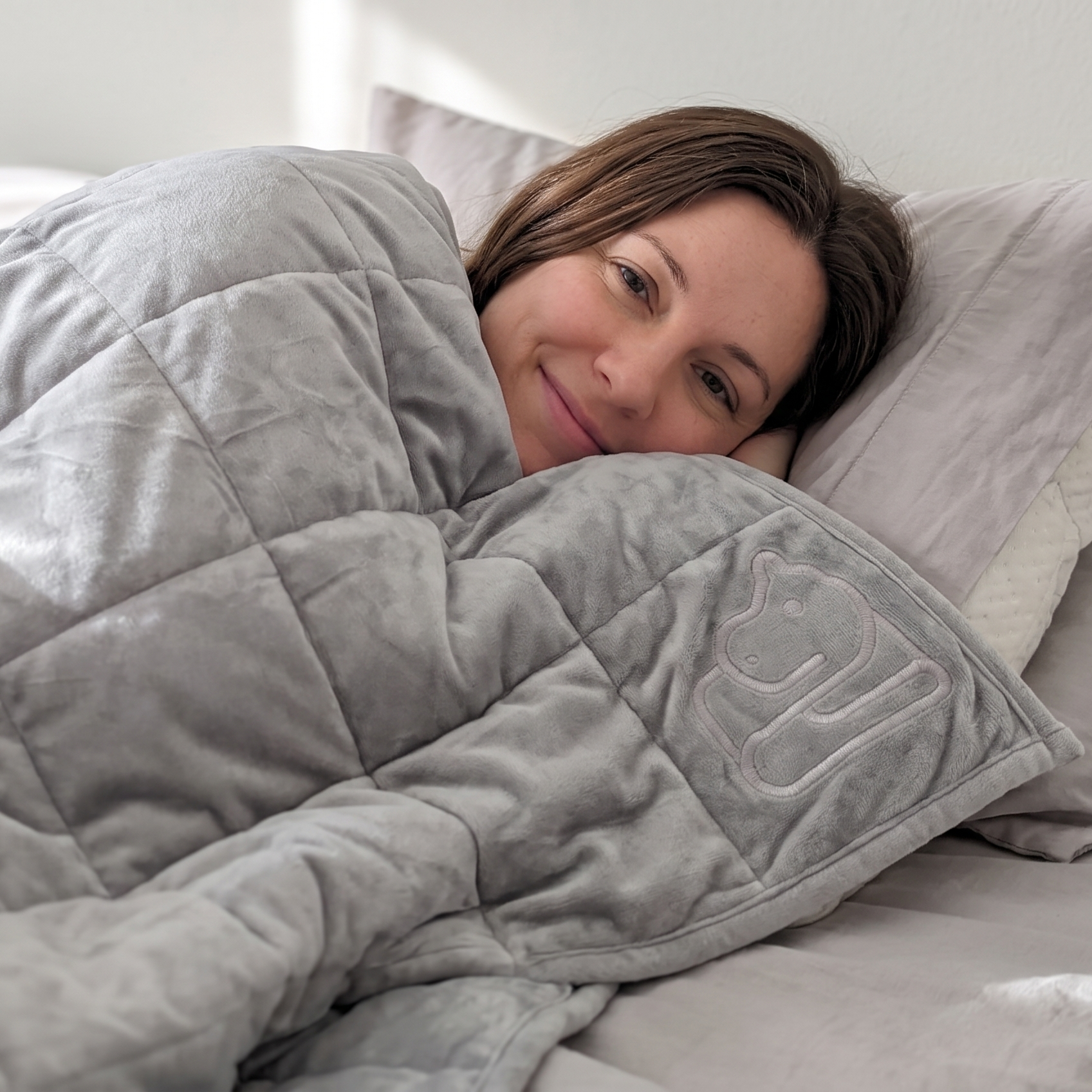 A woman lying in bed under a light gray weighted blanket with a matching pillow featuring an embroidered cat silhouette.