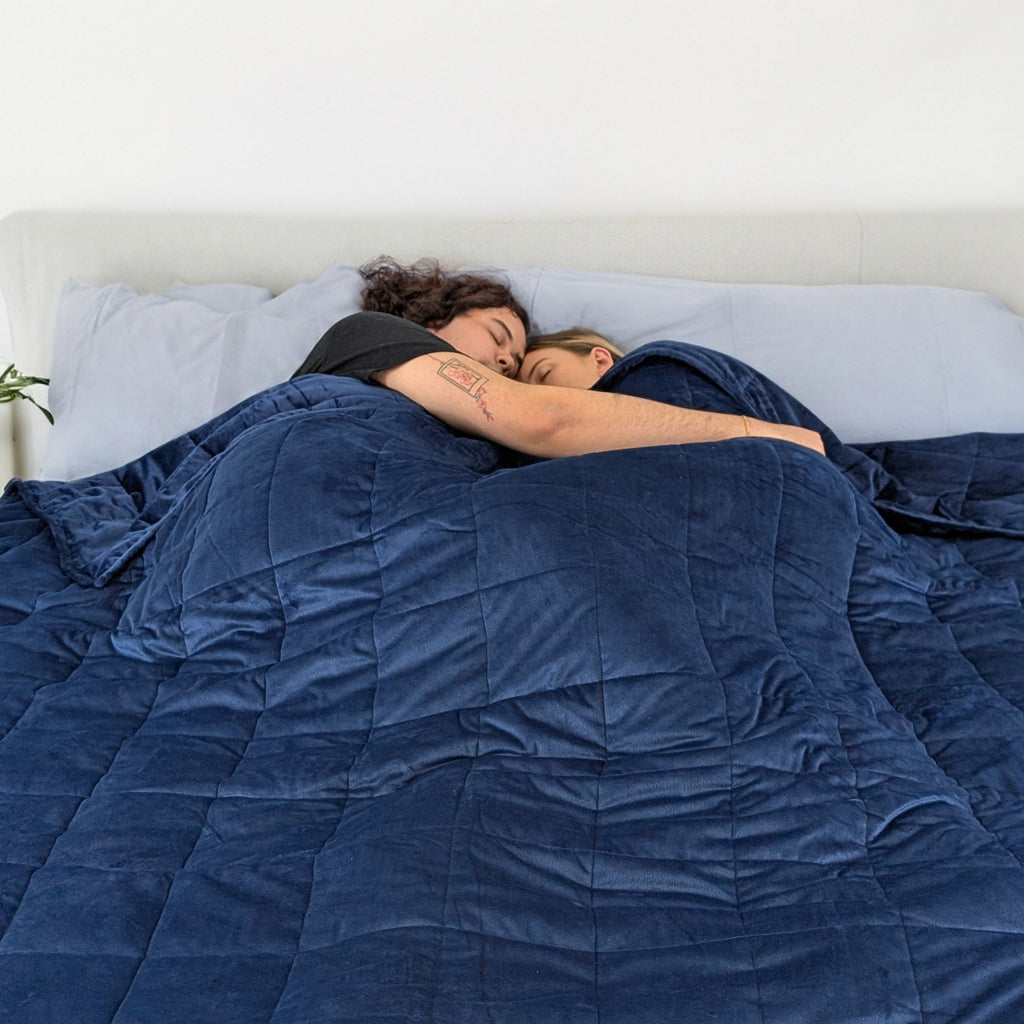 Two people under a blue weighted blanket on a bed.