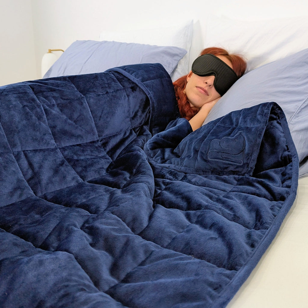 Person lying in bed under a blue weighted blanket with a sleep mask on.