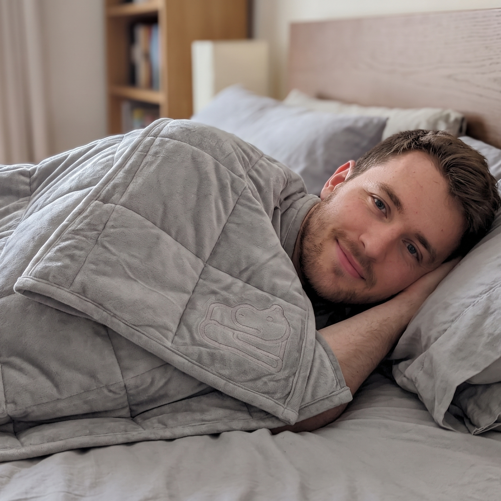 A man lying in bed under a light gray weighted blanket, looking at the camera with a soft, relaxed expression in a softly lit bedroom.
