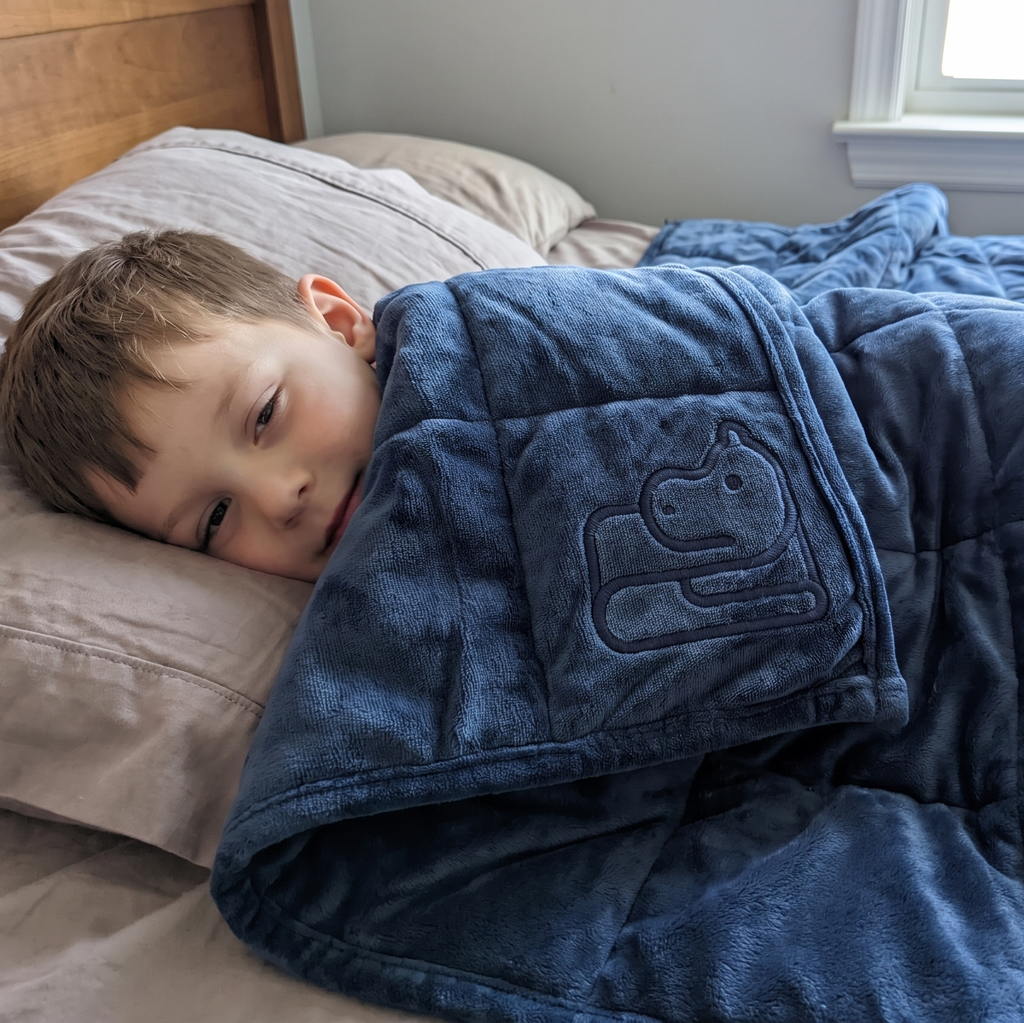 A young boy rests on a bed under a plush blue weighted blanket with an embroidered bear patch, in a bedroom setting.