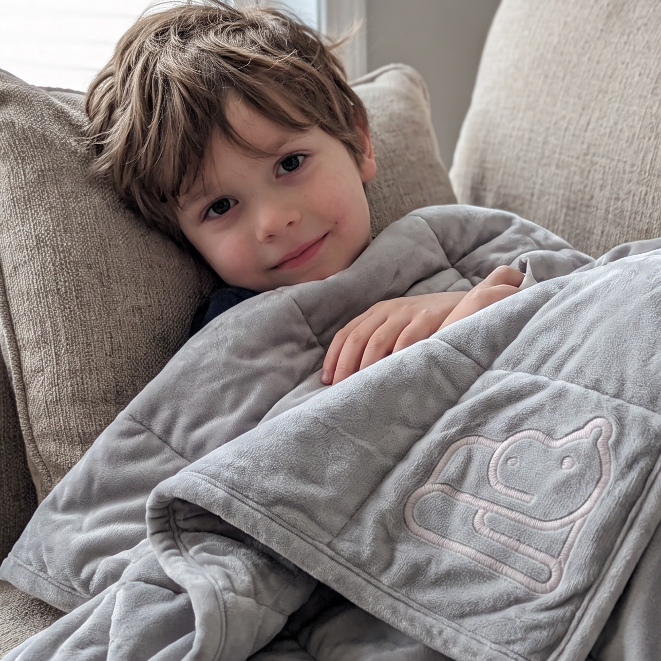 A young boy with light brown hair lies on a beige couch under a gray quilted weighted blanket with a silver embroidered hippo design.