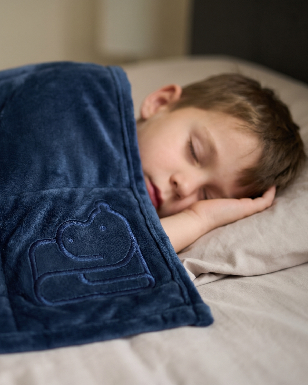 A child sleeping soundly under a dark navy blue quilted weighted blanket on a bed. The blanket features an embroidered outline of a hippo on the corner.